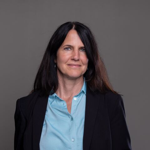 Una donna dai lunghi capelli scuri accenna un sorriso alla telecamera. Indossa un blazer nero e sotto una camicetta azzurra.