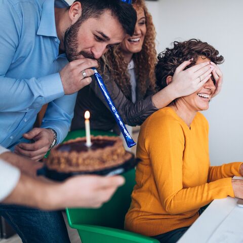 Una donna al computer viene sorpresa dai colleghi con una torta di compleanno, mentre un uomo suona un clacson e un'altra donna si copre gli occhi.