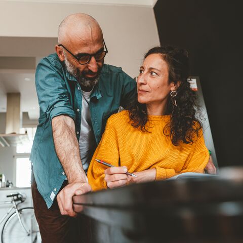 A man is leaning over and smiling at his wife who is sitting at the table and writing. In the background, a bicycle is propped up in the kitchen.