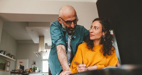 A man is leaning over and smiling at his wife who is sitting at the table and writing. In the background, a bicycle is propped up in the kitchen.