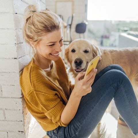 A smiling woman in a yellow sweater sits on the floor looking at her smartphone, while a Golden Retriever stands next to her.