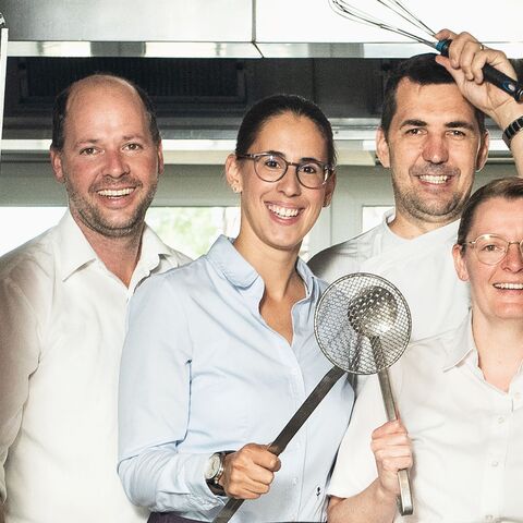 Un groupe de quatre cuisiniers souriants dans une cuisine, deux d'entre eux tenant dans leurs mains des ustensiles de cuisine tels qu'un fouet et une écumoire.