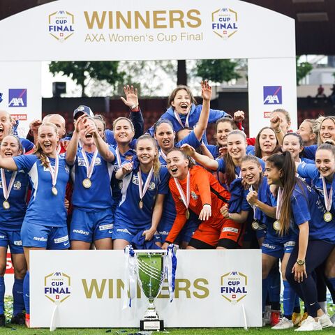 Eine Frauen-Fussballmannschaft feiert ihren Sieg nach dem AXA Women's Cup Finale, posierend mit Medaillen und einem Pokal unter einem "Winners"-Banner.