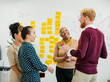 Quatre collaborateurs discutent devant un tableau blanc couvert de post-it jaunes.