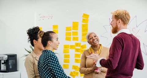 Quatre collaborateurs discutent devant un tableau blanc couvert de post-it jaunes.
