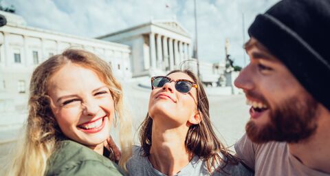 Three young, cheerful people laugh together outside in front of a historic building with columns and a statue in the background.