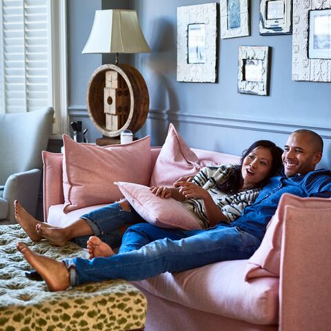 A smiling couple relaxes on a pink sofa in a cozy living room, surrounded by cushions and decorations.
