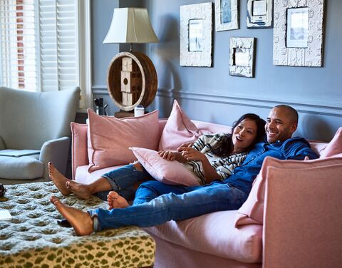 Ein lächelndes Paar liegt entspannt auf einem rosa Sofa in einem gemütlichen Wohnzimmer, umgeben von Kissen und Dekorationen.