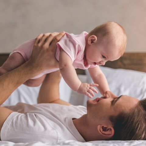 Un uomo è sdraiato a letto sorridente e tiene in braccio un bambino con una tutina rosa mentre si guardano e si toccano.