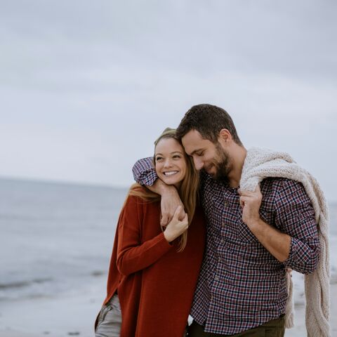 Una giovane coppia passeggia sulla spiaggia. L’uomo circonda con il braccio le spalle della donna sorridente.