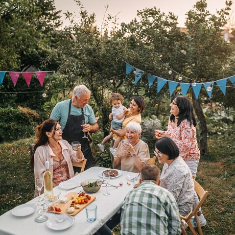 Une famille est assise et debout autour d'une table recouverte d'une nappe blanche dans le jardin. Ils rient et font la fête. Au-dessus d'eux, il y a une guirlande lumineuse et des triangles de fête bleus.