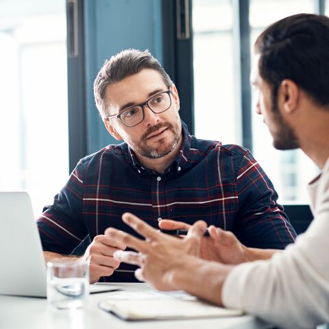 Zwei Männer sitzen an einem Tisch und diskutieren konzentriert, einer mit Brille und Laptop, der andere gestikuliert mit den Händen.