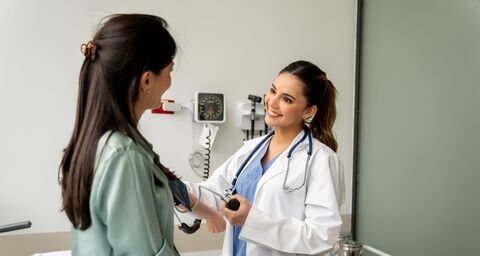 A smiling doctor is taking the blood pressure of a patient sitting on a bed.