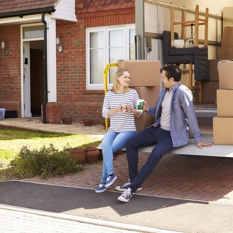 Un couple est assis sur la surface de chargement d’un camion de déménagement devant une maison en briques, entourés de cartons, en train de boire un café.