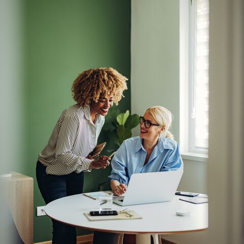 Two women laugh together and look at a laptop.
