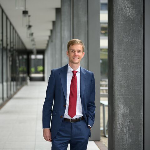 A man in a blue suit and red tie stands in a modern, glass office corridor and smiles at the camera.
