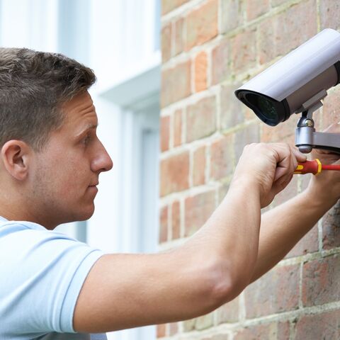Un homme installe une caméra de surveillance sur un mur de briques et serre une vis à l’aide d’un tournevis.