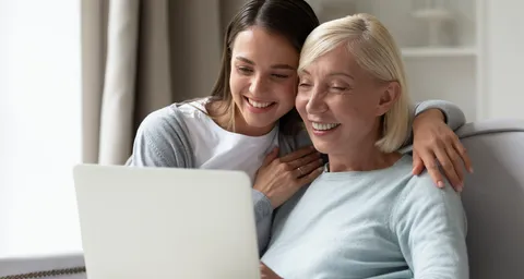 Lächelnde ältere Mutter und erwachsene Tochter sitzen entspannt auf dem Sofa im Wohnzimmer und schauen gemeinsam ein Video auf dem Laptop.