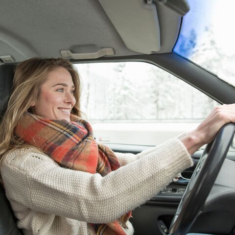 A smiling woman with long blonde hair and a checkered scarf drives a car on a snowy day looking forward.