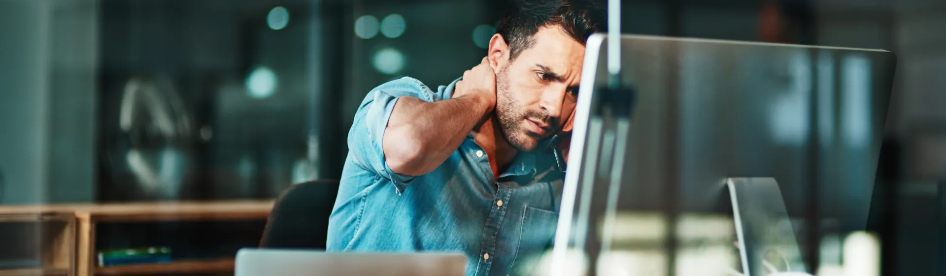 An exhausted man sits at his desk in front of his laptop, holding his forehead.