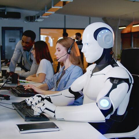 Bild zeigt eine Bürosituation in der ein humanoider Roboter neben menschlichen Angestellten am Bildschirm sitzt und arbeitet.