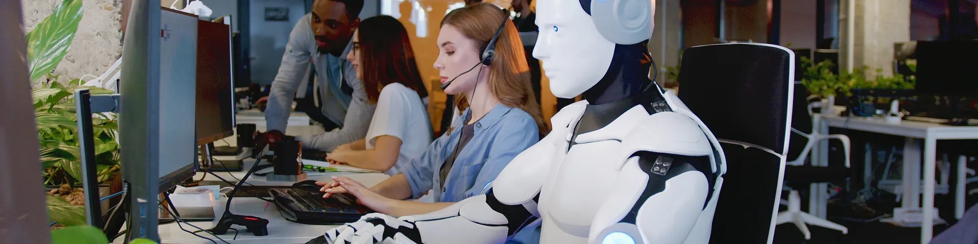 Bild zeigt eine Bürosituation in der ein humanoider Roboter neben menschlichen Angestellten am Bildschirm sitzt und arbeitet.