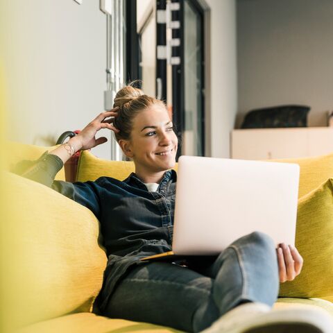 Eine Frau sitzt entspannt auf einem gelben Sofa und arbeitet lächelnd an einem Laptop, umgeben von einem modernen, gemütlichen Raum.