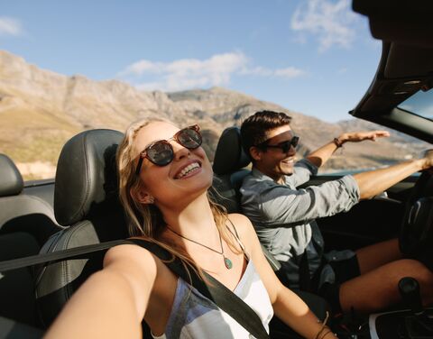 Par une journée ensoleillée, un couple souriant traverse un paysage montagneux dans une voiture décapotable, tous deux portant des lunettes de soleil.