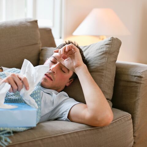 Ein Mann liegt zuhause krank auf dem Sofa. Er nimmt sich ein Kleenex aus der Box und hält seine Hand an die Stirne.