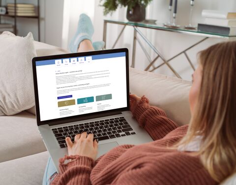 Eine Frau sitzt zuhause gemütlich auf dem Sofa und hat ihren Laptop auf dem Schoss. 