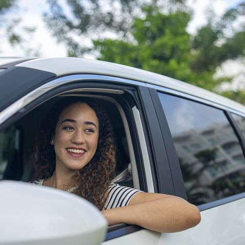 Junge Frau sitzt lächelnd am Steuer eines Autos und schaut aus dem geöffneten Fenster.