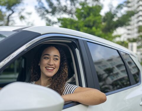 Junge Frau sitzt lächelnd am Steuer eines Autos und schaut aus dem geöffneten Fenster.