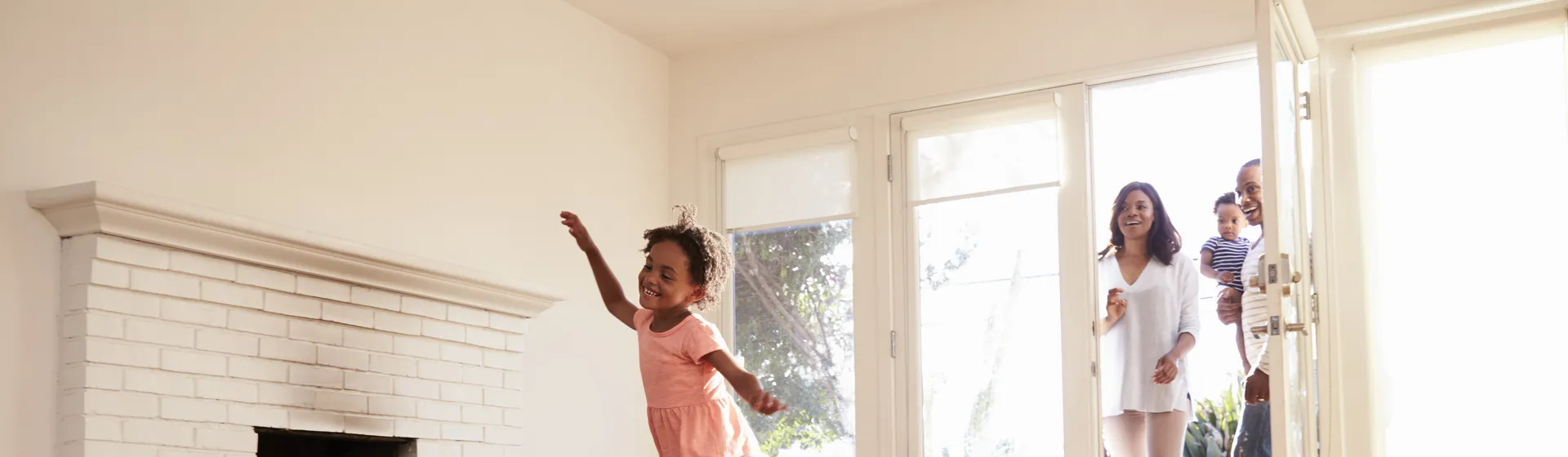 A little girl runs joyfully through an empty living room with a fireplace, while three smiling adults come in through the open door.