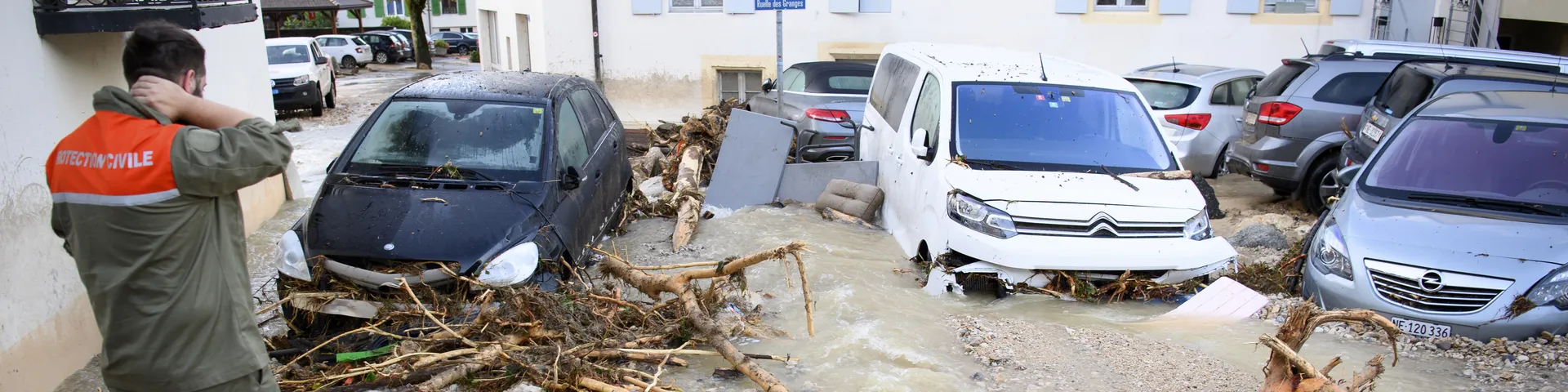 Ein Strassenabschnitt ist überflutet, Autos stehen im Wasser, und Trümmer sind verstreut. Ein Mann in Uniform betrachtet die Szene.