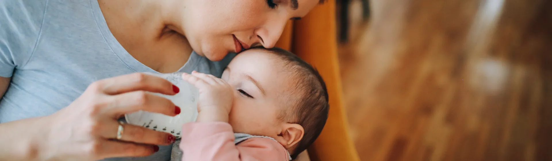 Une femme nourrit affectueusement un bébé avec un biberon et embrasse son front, assise sur une chaise dans une pièce au sol en bois.