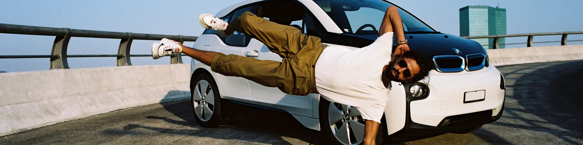 Un homme vêtu d’un t-shirt blanc et d’un pantalon marron fait un arrêt devant une voiture blanche sur un pont à la lumière du jour.