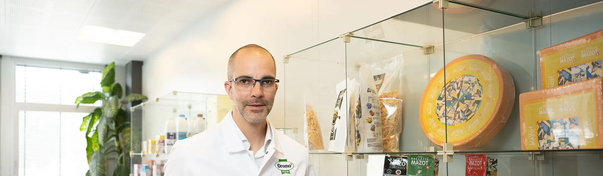 Un uomo in camice bianco e occhiali è in piedi in uno showroom. Sugli scaffali di vetro sono esposti diversi prodotti caseari.