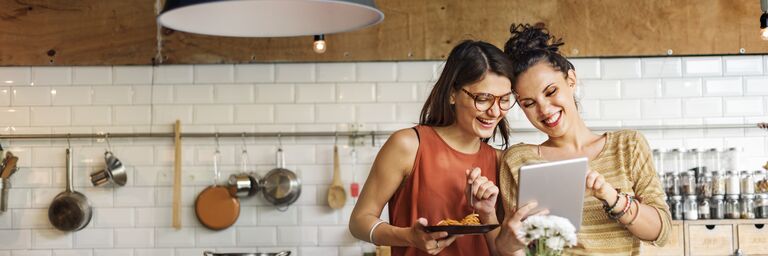 Zwei lachende Frauen stehen in einer modernen Küche, essen Pasta und schauen gemeinsam auf ein Tablet