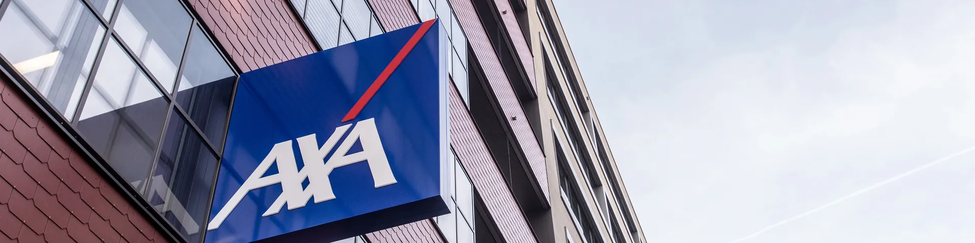 Ein modernes Bürogebäude mit roten Ziegeln und grossen Fenstern, an dem ein blaues AXA-Logo mit weisser Schrift und rotem Streifen angebracht ist.