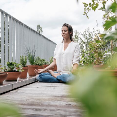 Une femme médite paisiblement sur un balcon, entourée de nombreuses plantes vertes en pots, et profite du calme.