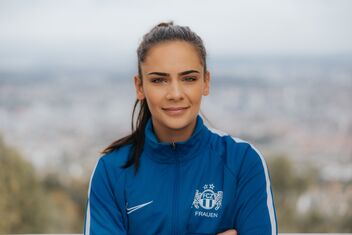 Une femme aux cheveux foncés attachés en arrière porte une veste de sport bleue avec le logo du FCZ Femmes et se tient à l'extérieur devant un arrière-plan flou.