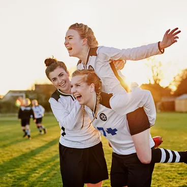 Trois joueuses de football en maillot blanc font la fête sur un terrain au coucher du soleil, l'une portant l'autre sur son dos, tout le monde rit.