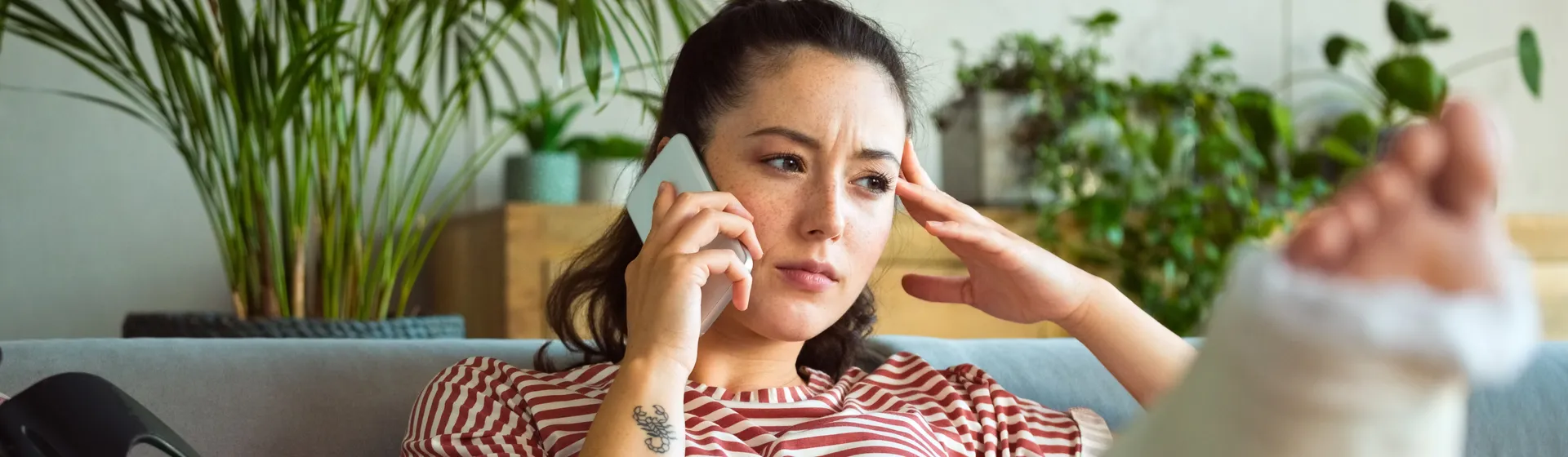 A woman with a plastered leg sits on a sofa talking on the phone, surrounded by plants in the background. She looks worried.