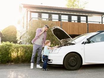 Un homme téléphone à côté d’une voiture en stationnement, capot moteur ouvert, tandis qu’un enfant regarde le compartiment moteur avec curiosité.
