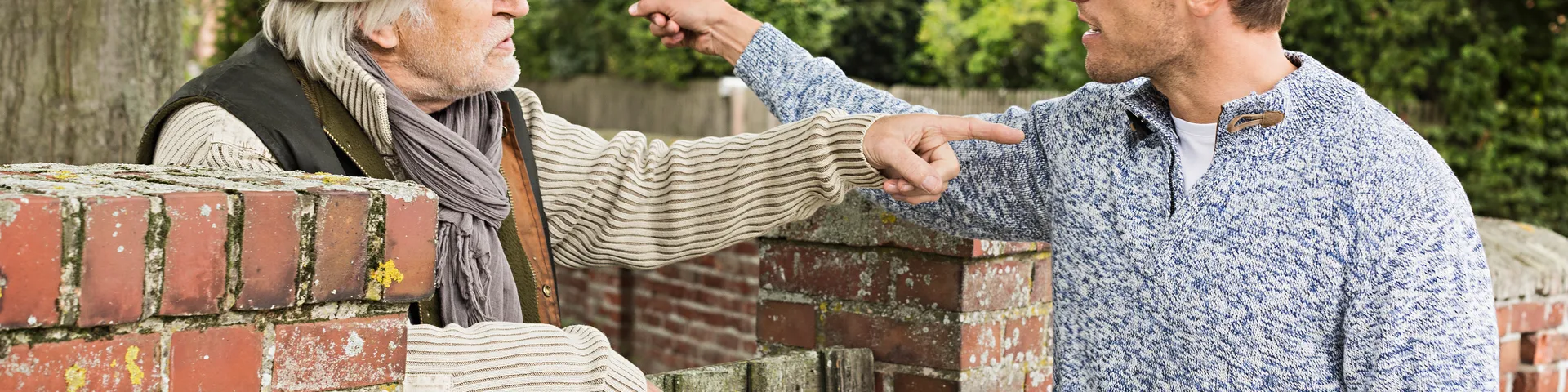 Un uomo anziano e un uomo più giovane stanno davanti a un muro di mattoni e si puntano gesticolando l’un l’altro mentre evidentemente litigano.