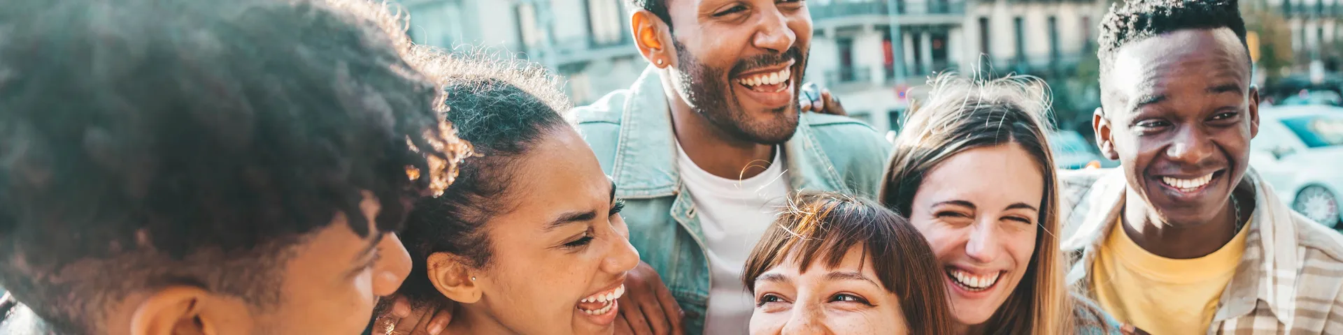 A group of young people of different genders and backgrounds laugh together outdoors in front of a building in a city.