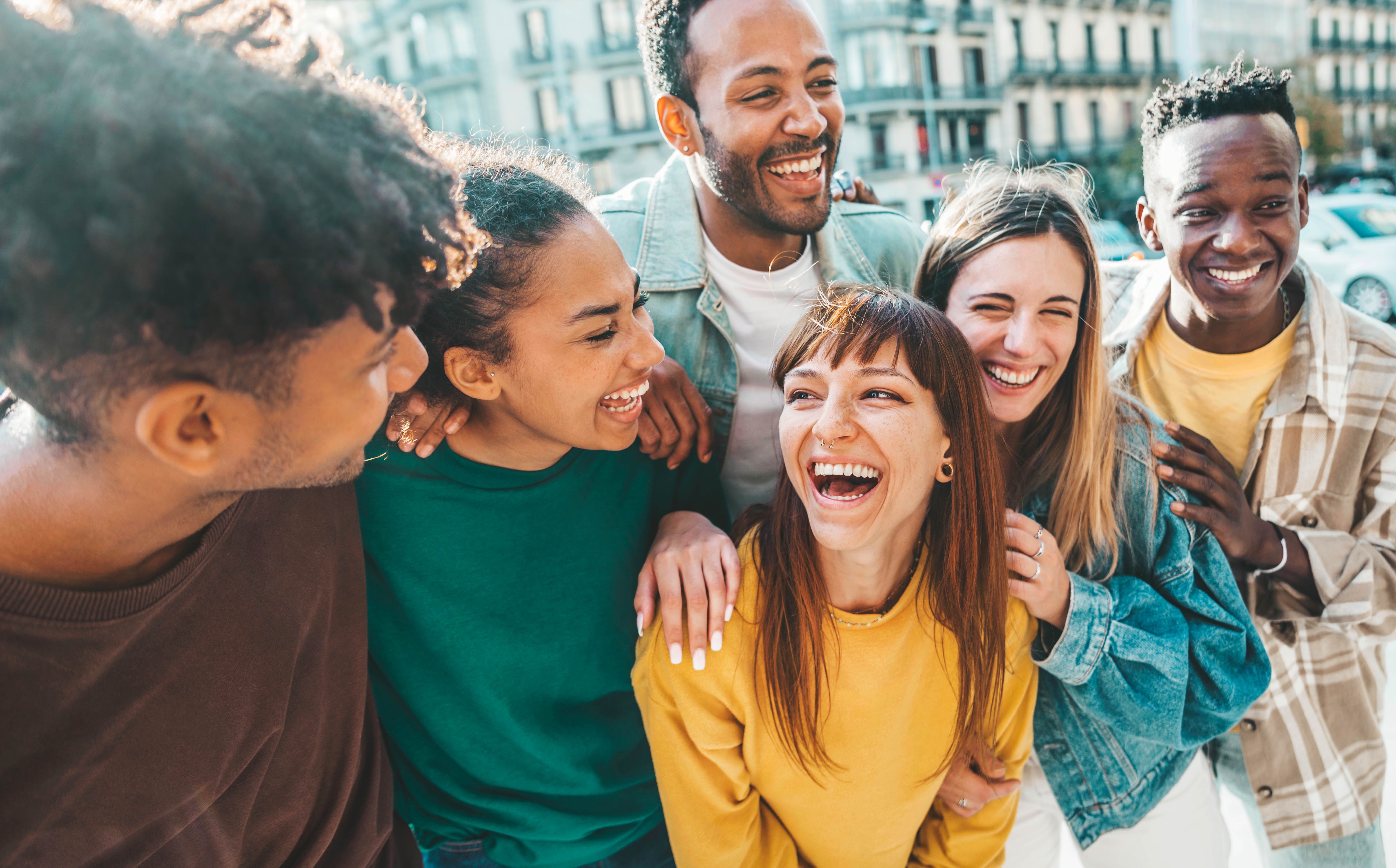 A group of young people of different genders and backgrounds laugh together outdoors in front of a building in a city.
