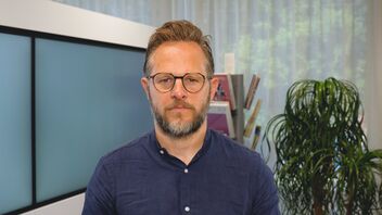 Ein Mann mit Brille, Bart und blauem Hemd steht in einem Eingangsbereich eines Unternehmens.