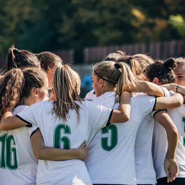 Un groupe de joueuses de football en maillot blanc s'embrasse en cercle sur un terrain vert, prêtes à jouer.