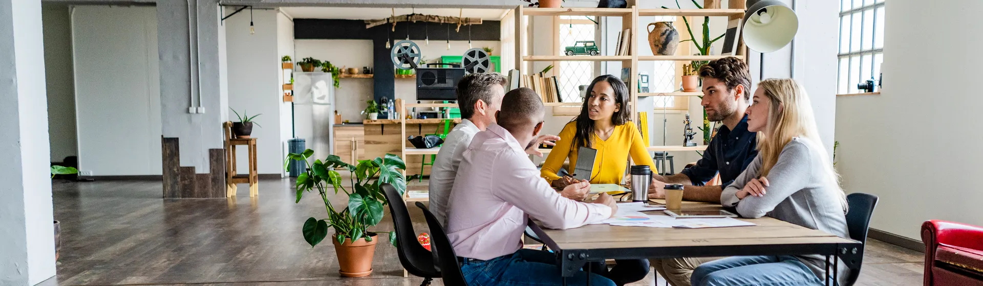 Fünf Personen sitzen an einem Tisch in einem modernen, offenen Büro mit Pflanzen und Regalen im Hintergrund und diskutieren angeregt.
